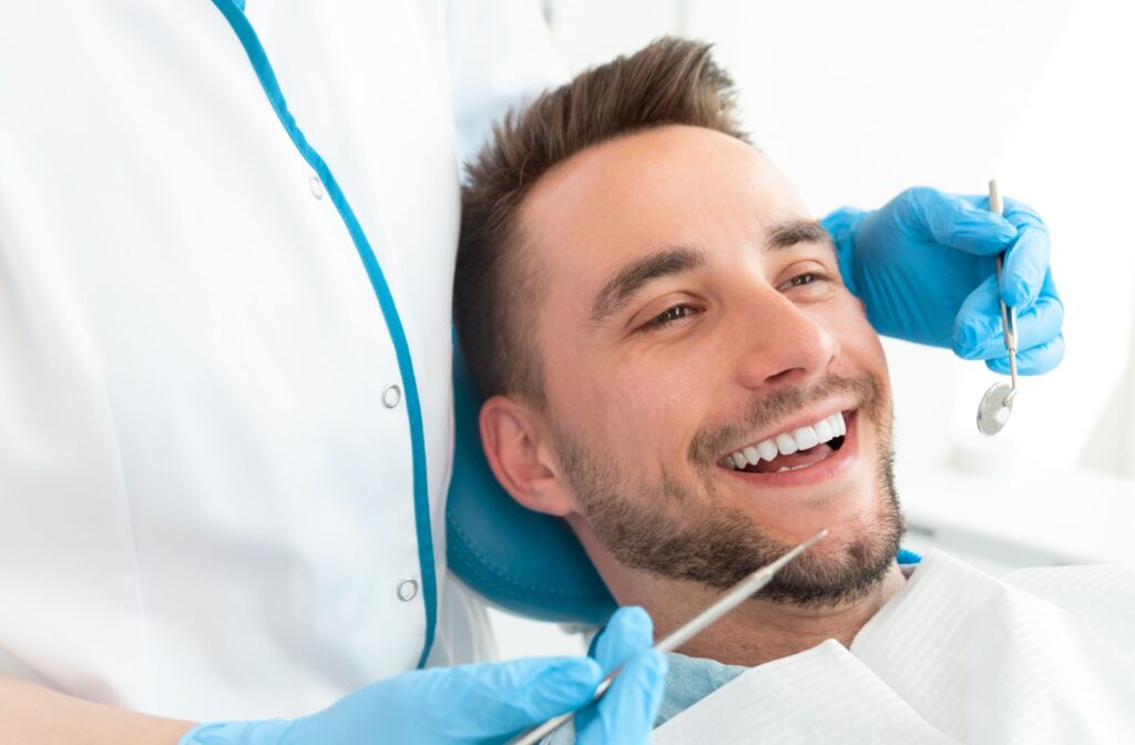 Smiling adult man in dental chair during routine checkup, with a dental hygienist holding instruments.