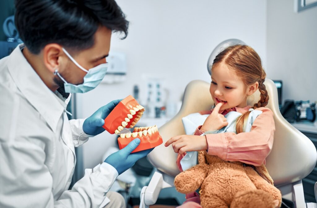 Young girl at the dentist holding a stuffed animal while learning about teeth from a dental professional using a model.