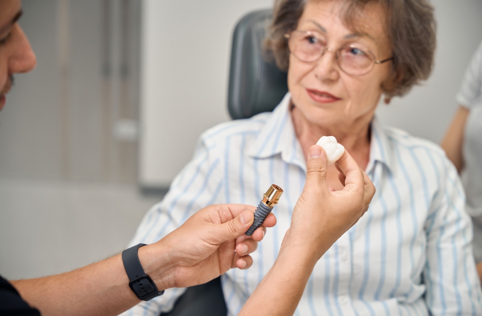 A senior patient talking to a dentist about a tooth implant while the dentist holds a 3D model of a tooth implant